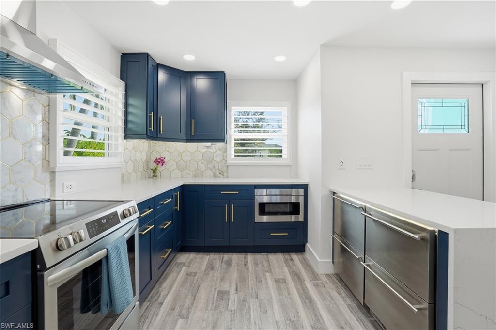 Kitchen featuring stainless steel appliances, a peninsula, wall chimney exhaust hood, blue cabinets, and recessed lighting - Virtually Edited Image