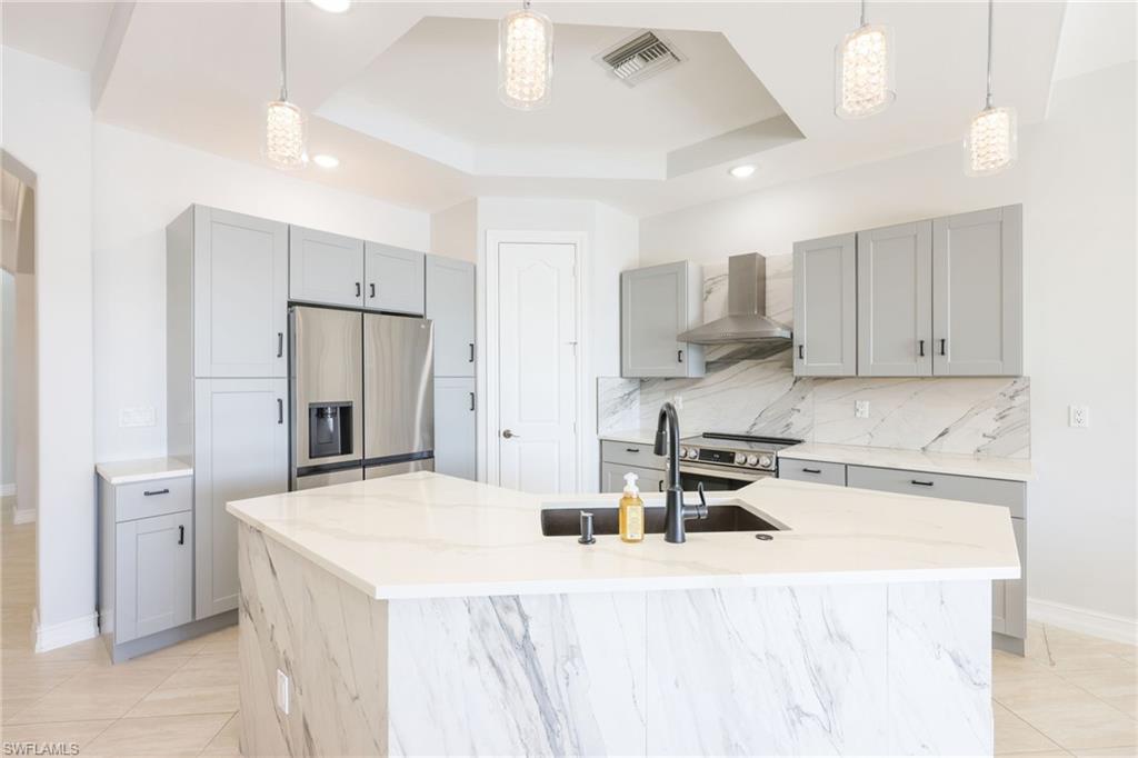 Kitchen featuring a central island with a waterfall edge, light gray cabinetry, stainless steel appliances, marble-patterned countertops, and a matching backsplash