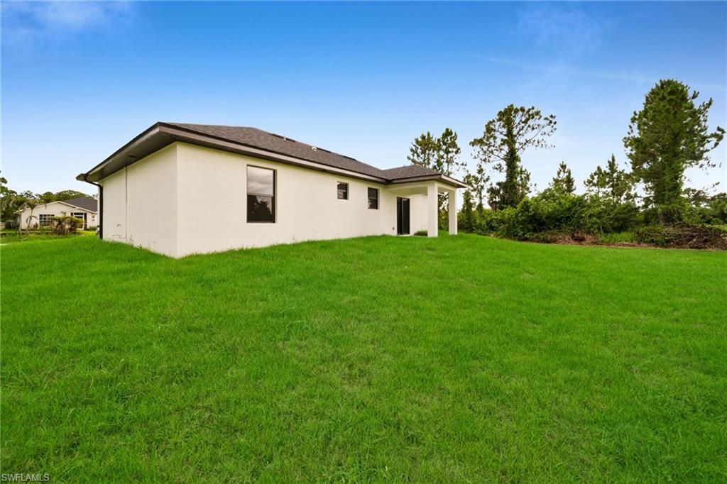 Expansive rear yard featuring a verdant lawn, a white stucco exterior, dark-framed windows, a dark shingle roof, and a covered patio area