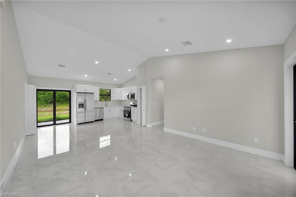 Open-concept living area featuring a vaulted ceiling with recessed lighting, large format tile flooring, and a contemporary kitchen with white cabinetry, stainless steel appliances, and a window overlooking exterior greenery