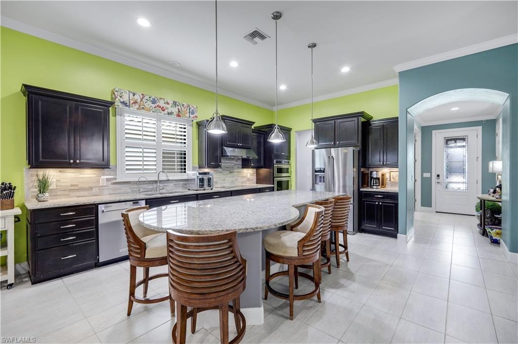 Spacious kitchen featuring an expansive oval island with seating, dark wood-finish cabinetry, stainless steel appliances, light-toned backsplash, and recessed lighting