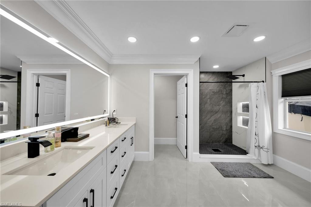 Spacious bathroom featuring a dual vanity with undermount sinks and black hardware, an illuminated mirror, a walk-in shower with dark stone-look tile, and recessed lighting