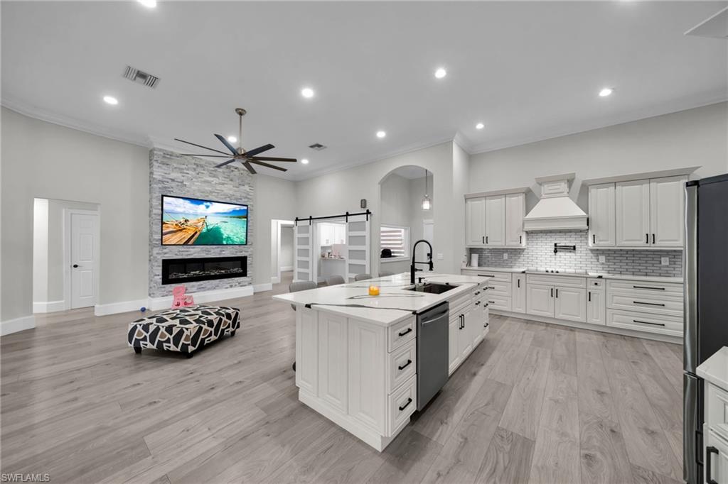Open-concept living space featuring wood-finish flooring, a kitchen island with integrated sink, a stacked stone fireplace surround, a modern ceiling fan, and white cabinetry with dark hardware