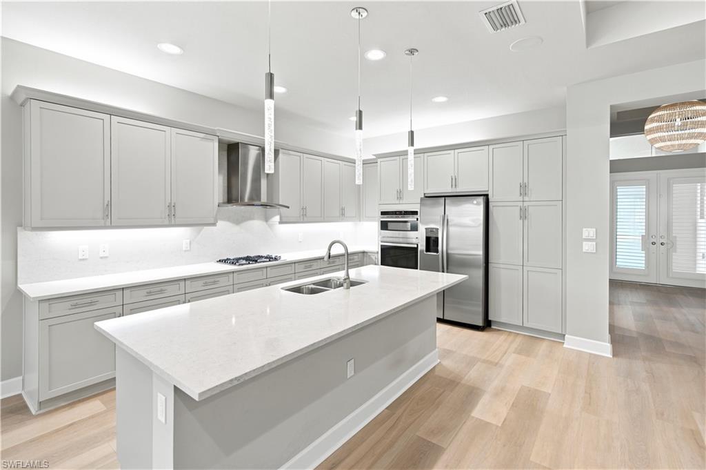 Contemporary kitchen featuring light gray cabinetry, stainless steel appliances, a large center island with a double sink, wood-finish flooring, and pendant lighting