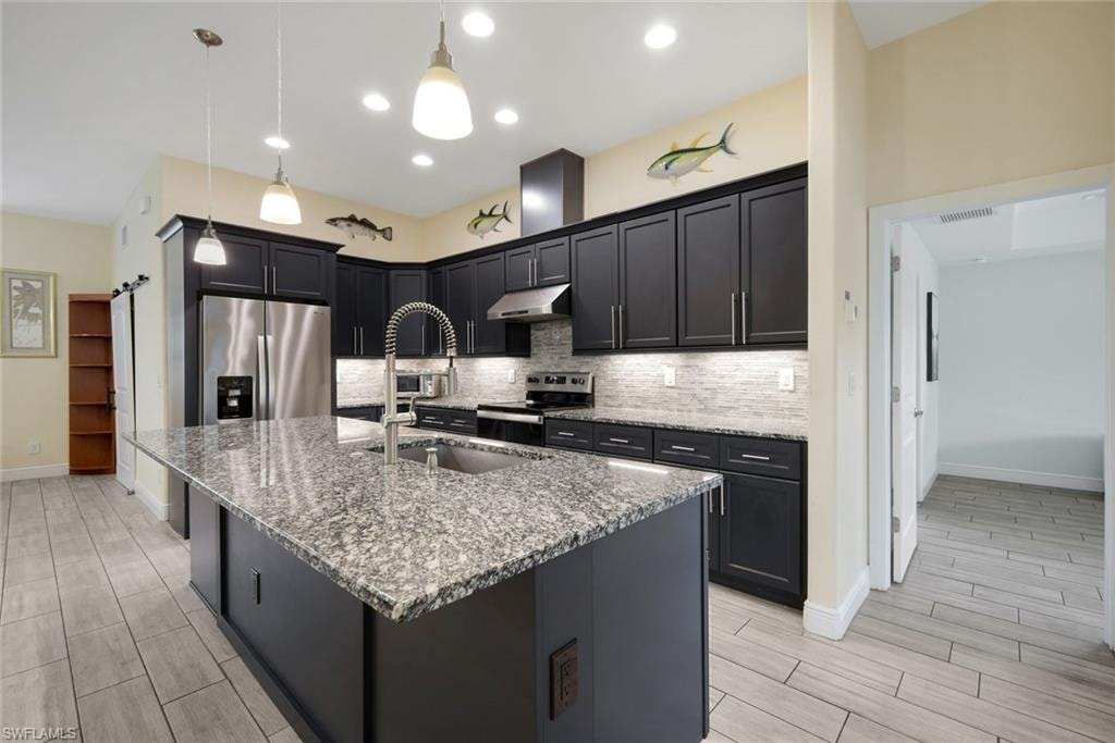 Kitchen featuring a large island with a speckled countertop, dark cabinetry, stainless steel appliances, and a tiled backsplash
