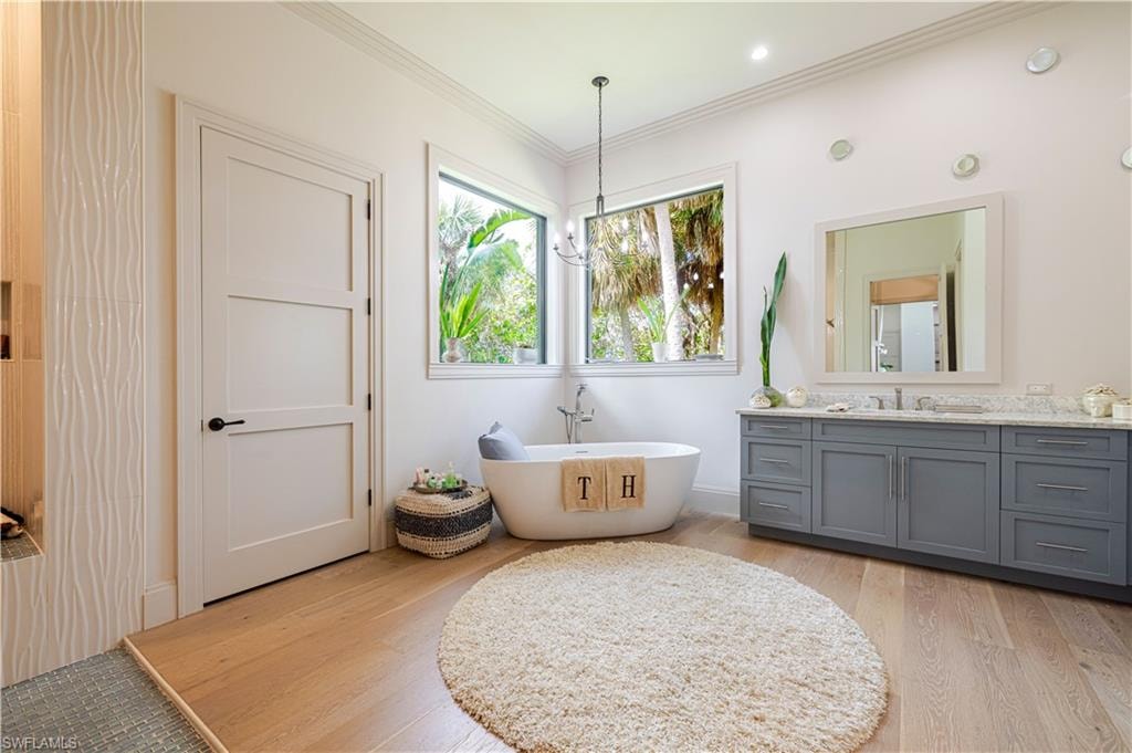 Freestanding soaking tub with a contemporary pendant fixture, large windows, light wood-finish flooring, and a double vanity with a stone countertop