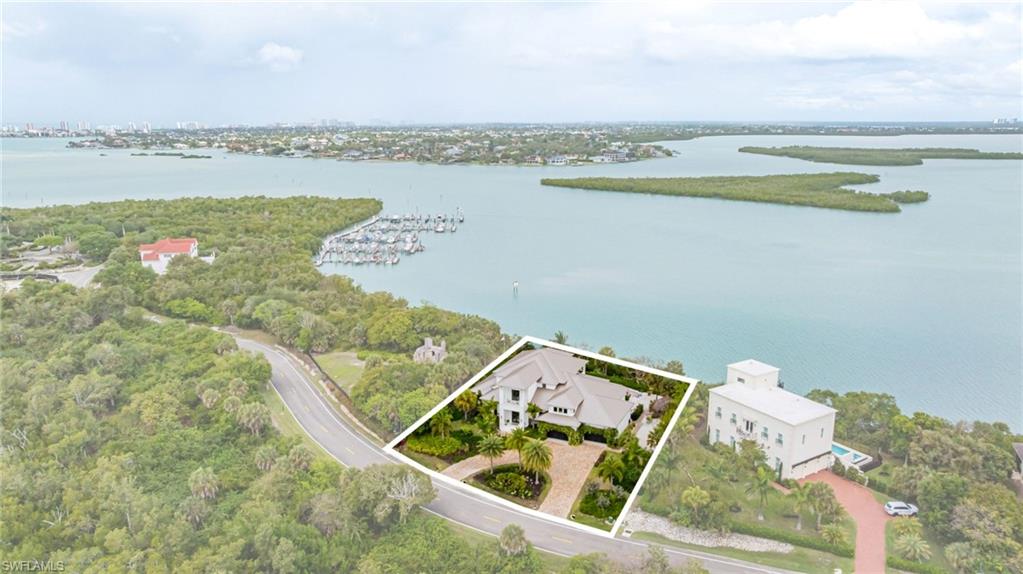 Expansive waterfront property featuring a large residence with a multi-gabled roofline, a palm-lined paved driveway, lush surrounding greenery, and direct water access