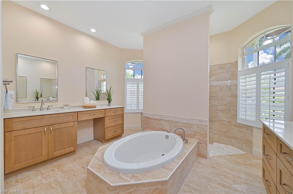 Spacious bathroom featuring a built-in soaking tub with surrounding tiled deck, dual wood-finish vanities with light countertops, arched windows with plantation shutters, a tiled shower enclosure, and recessed lighting