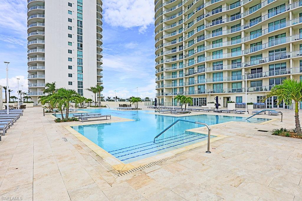 Resort-style pool deck with multiple swimming areas, expansive lounge seating, mature palm trees, and light-colored stone paving, flanked by two high-rise buildings featuring extensive private balconies