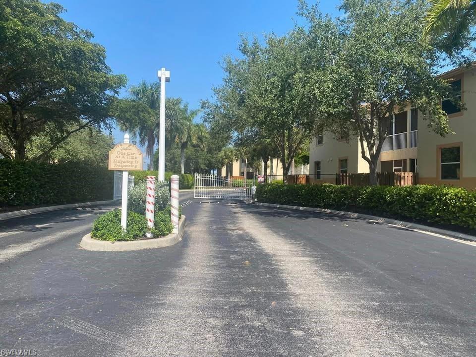 Gated community entrance featuring a paved driveway, lush perimeter landscaping, established trees, and multi-story residential buildings in the background