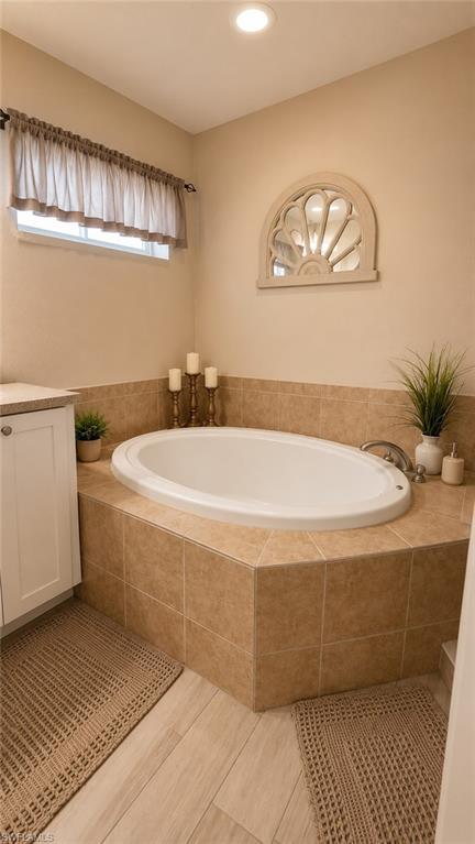 Built-in soaking tub with tiled surround, light wood-finish flooring, recessed lighting, and a white vanity cabinet with a light-toned countertop