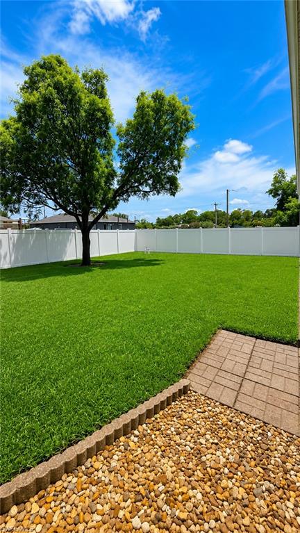 Fenced backyard with expansive artificial turf, a mature shade tree, a paved patio section, and decorative river rock landscaping
