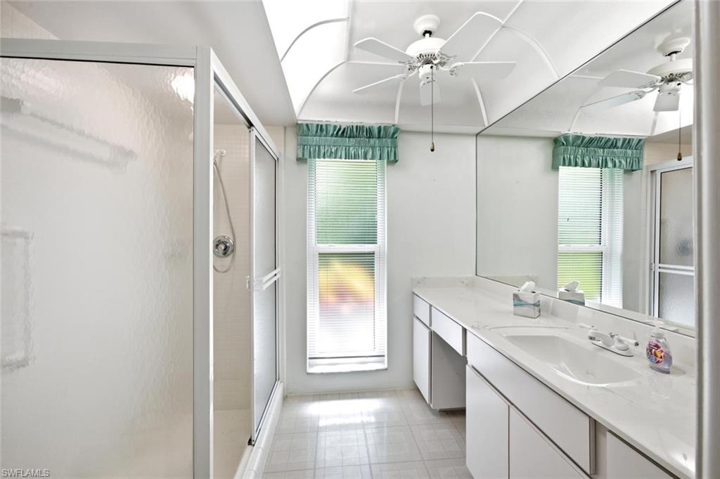 Bathroom featuring a shower enclosure with a sliding door, a dual-sink vanity with ample countertop space, a wall-mounted mirror, a ceiling fan, and a skylight