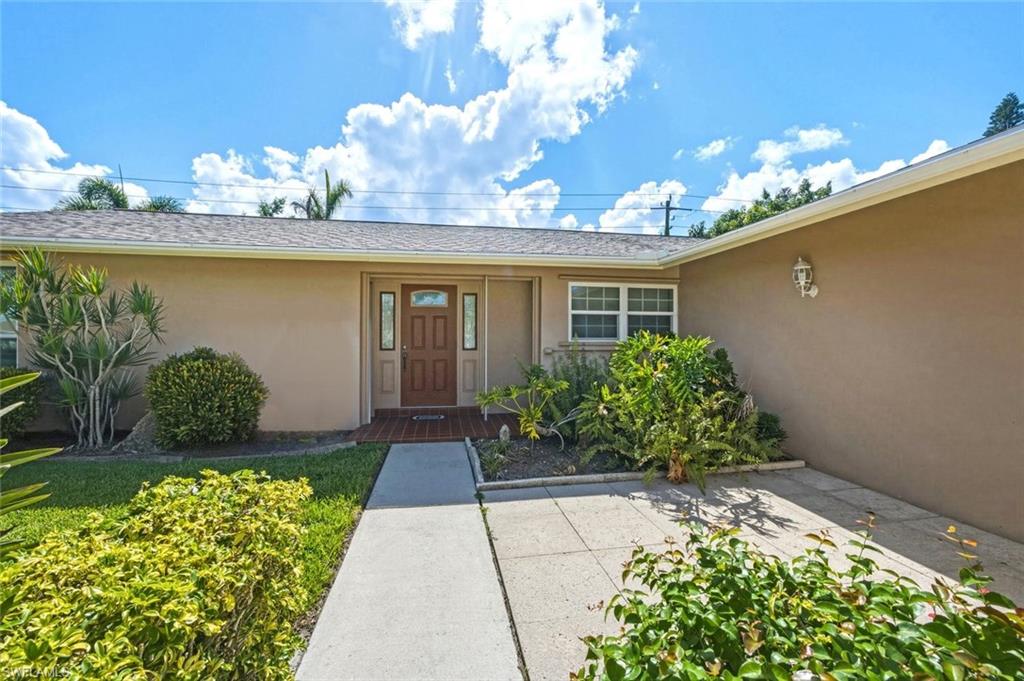 Stucco exterior featuring a wood-paneled entry door with sidelight windows, a covered entryway with brick-finish flooring, a concrete sidewalk, and mature landscaping