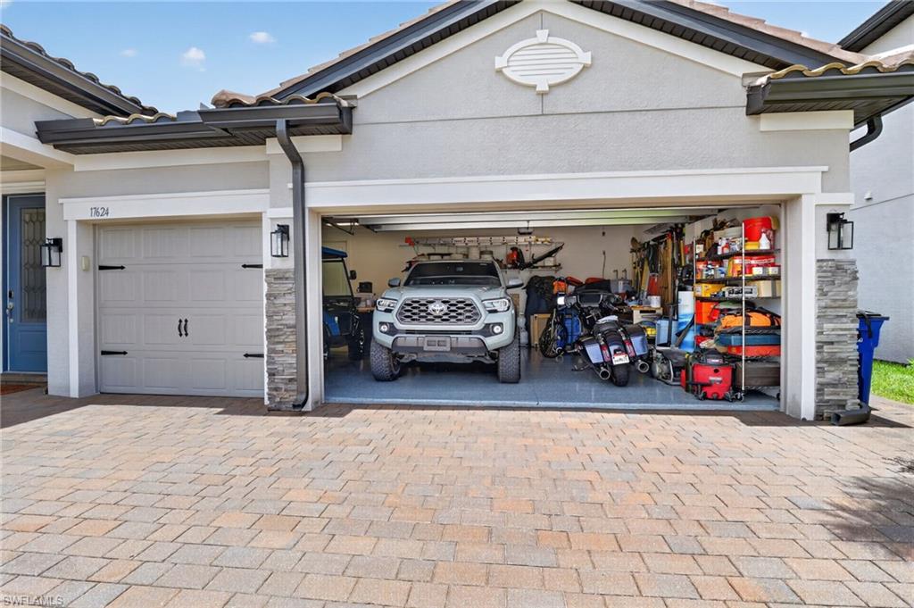 Two-car garage with a paver driveway, gray stucco exterior, stone accent columns, tiled roof, and a painted front door with sconce lighting