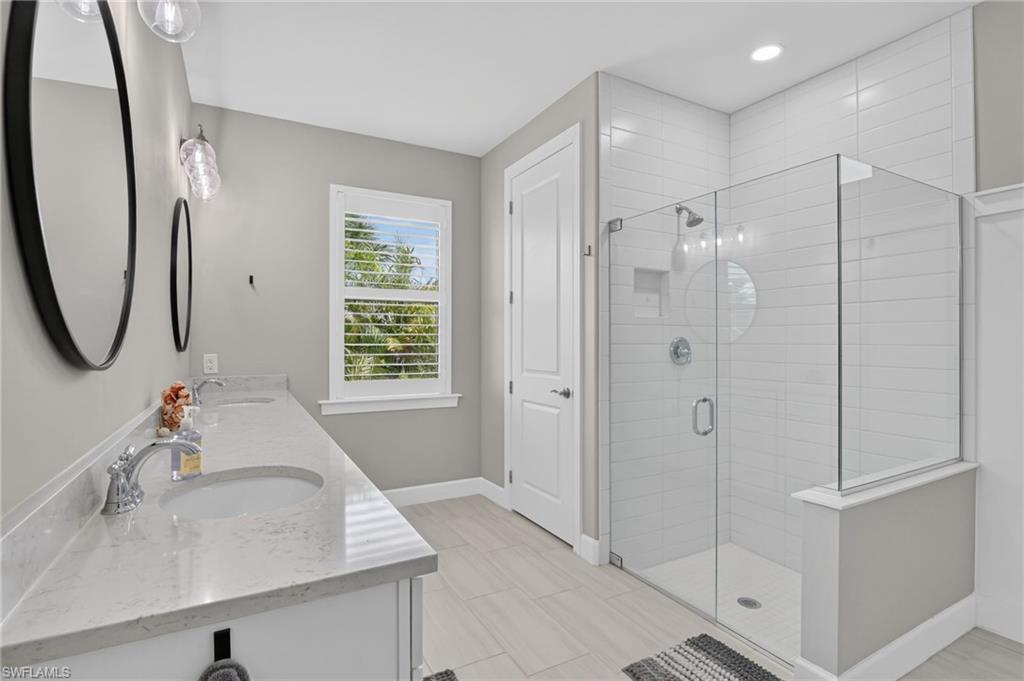Modern bathroom featuring a dual vanity with undermount sinks and light-toned countertops, a glass-enclosed shower with horizontal tile surround, a window with louvered blinds, and light wood-finish flooring