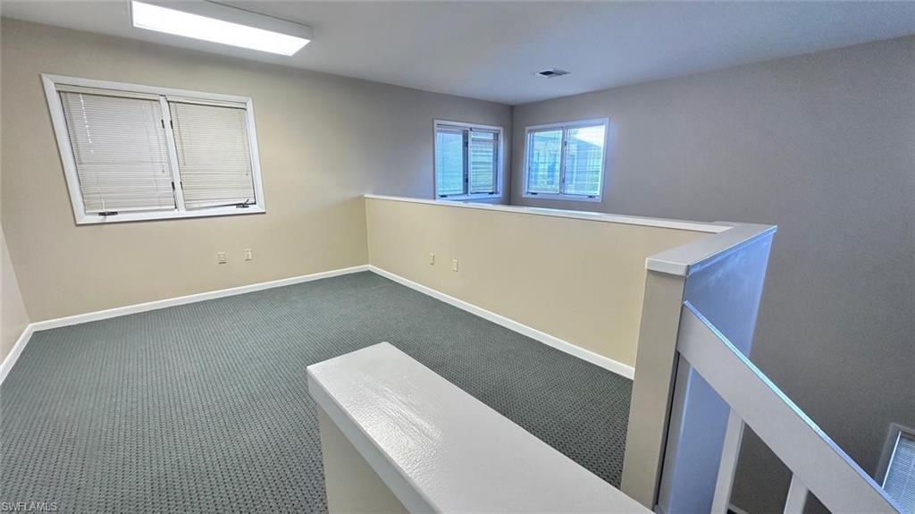 Carpeted loft area featuring a white railing, two windows with blinds, a rectangular fluorescent light fixture, and neutral-toned walls