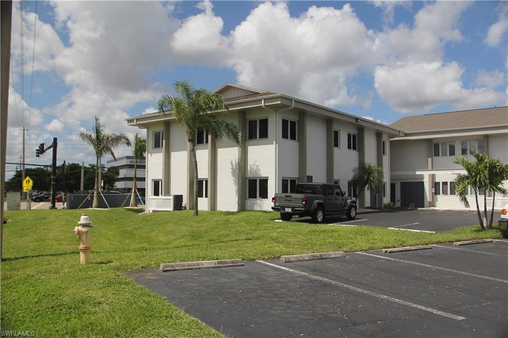 Two-story commercial building featuring a light-colored stucco exterior, multiple window openings, and an asphalt parking lot with marked spaces