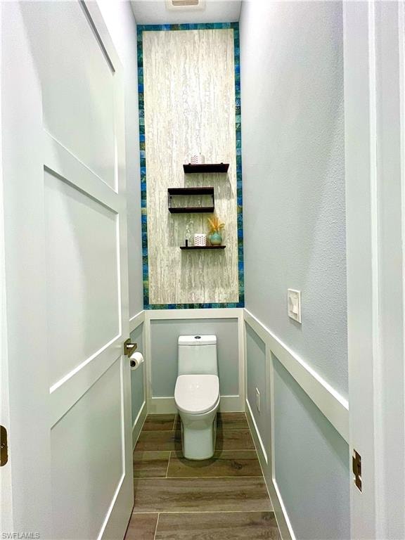 Powder room featuring wood-finish tile flooring, wainscoting, a contemporary toilet, and a tiled accent wall with floating shelves