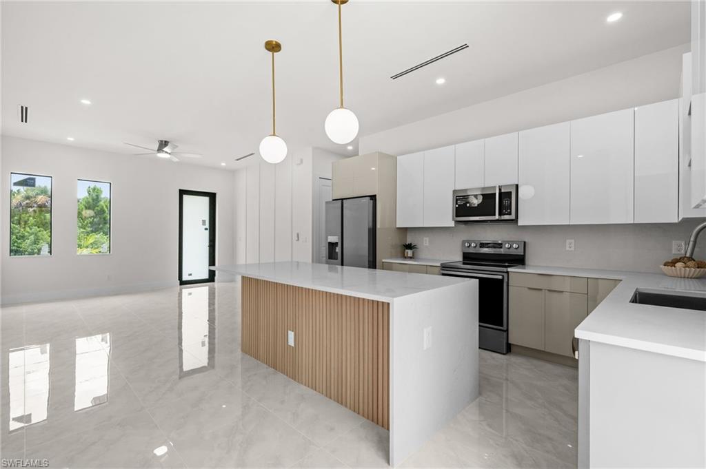 Open-concept kitchen and living area featuring a large center island with wood-slat detailing, white gloss cabinetry, integrated stainless steel appliances, and sleek tile flooring throughout