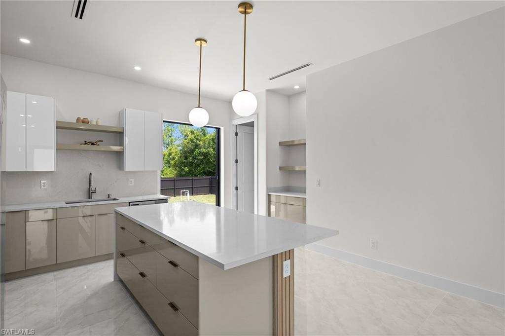 Modern kitchen featuring an expansive island with a light-toned countertop, sleek cabinetry with varying finishes, integrated sink with a matte black faucet, open shelving, and a large window overlooking a fenced outdoor area