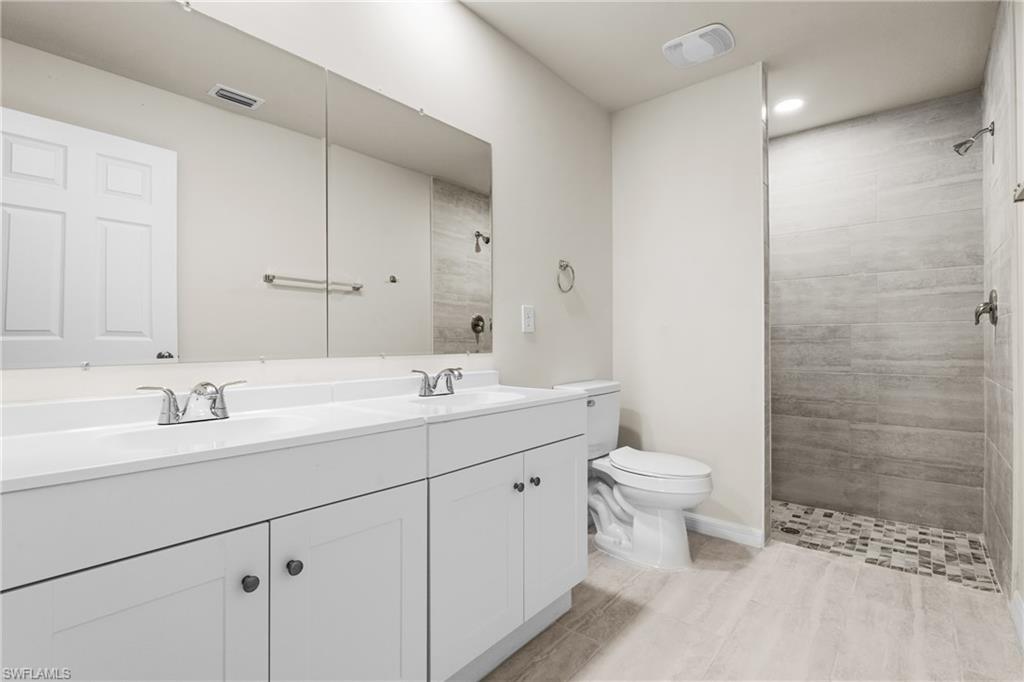 Bathroom featuring a double vanity with white cabinetry, a large wall-mounted mirror, a walk-in shower with textured tile, a mosaic shower floor, and light-toned floor tile
