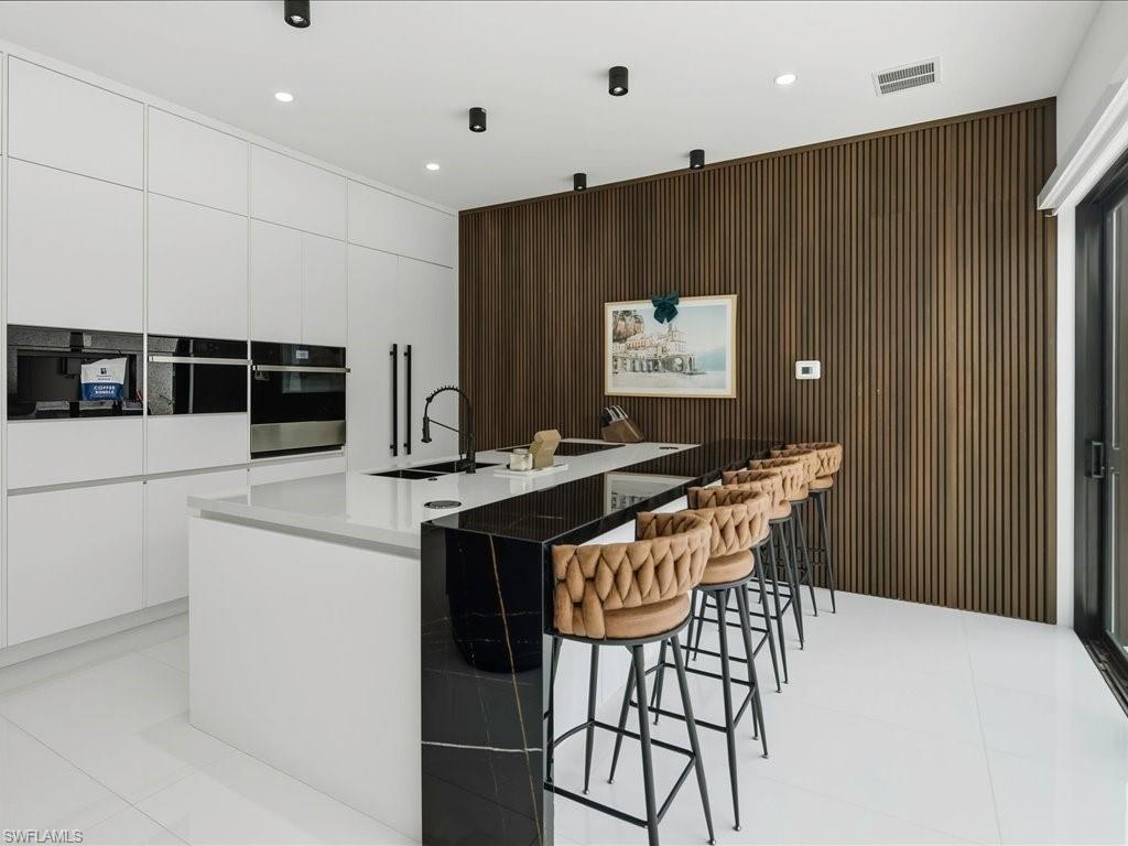 Contemporary kitchen featuring a white monochrome cabinetry wall with integrated appliances, a multi-level island with a black and white waterfall countertop, white floor tiling, and a textured wood-finish accent wall