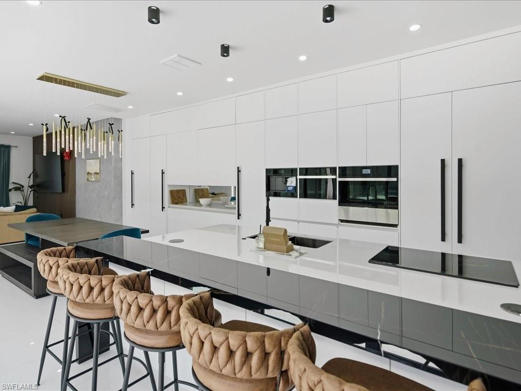Spacious kitchen featuring a large central island with integrated cooktop and sink, sleek white cabinetry with long black pull handles, and built-in stainless steel appliances