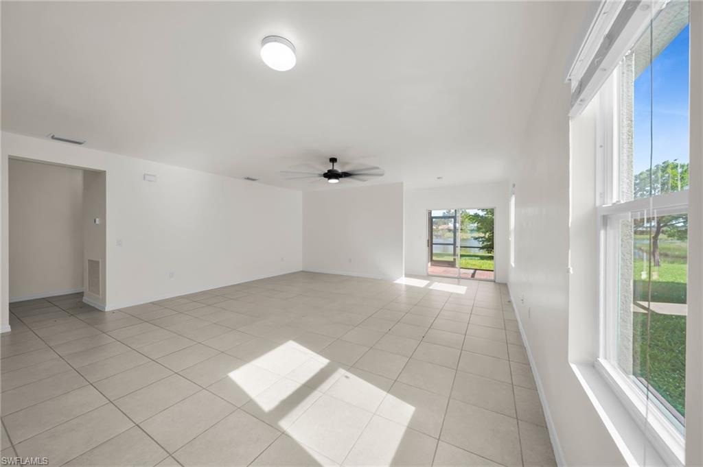 Spacious living area featuring light tile flooring, white walls, multiple windows, a ceiling fan, and sliding glass doors opening to an exterior area with a body of water