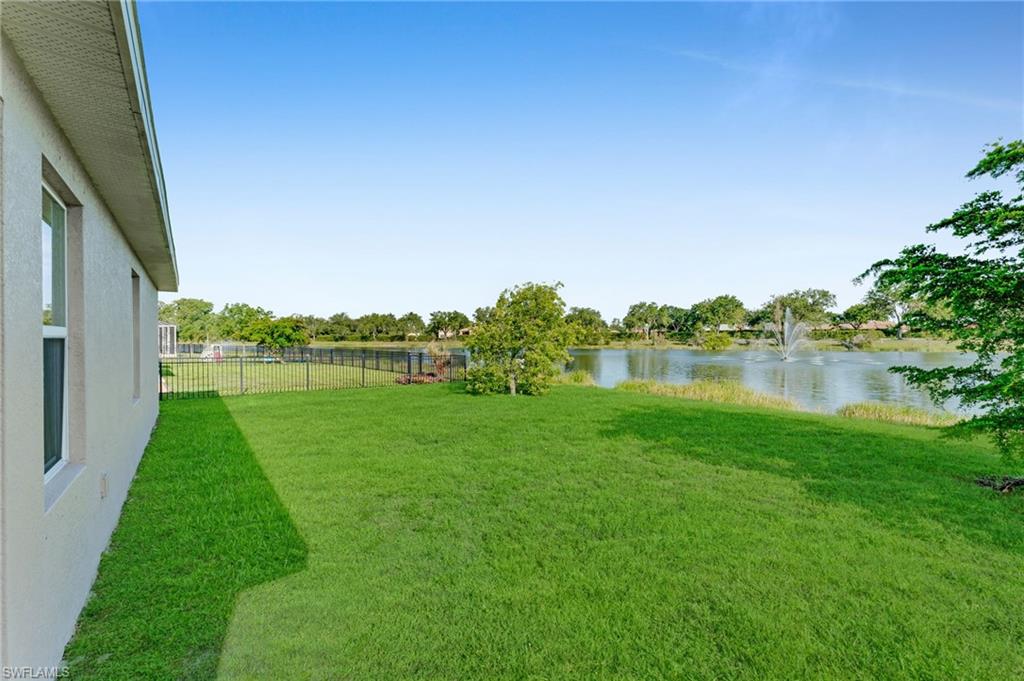 Grassy backyard featuring a black metal fence, mature landscaping, and water frontage with a fountain