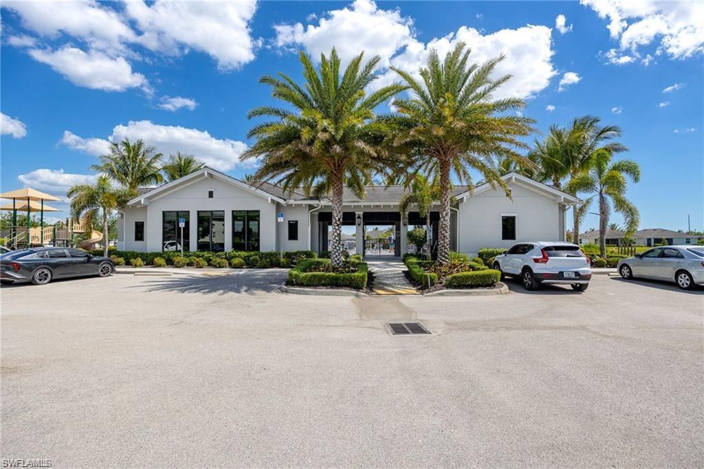 Modern clubhouse exterior featuring a light gray facade, dark framed windows, lush landscaping, mature palm trees, and a paved parking area