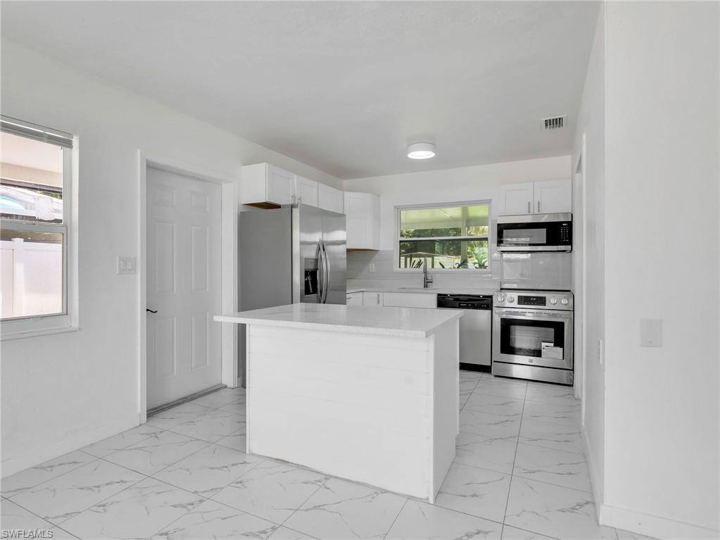 Modern kitchen featuring white shaker cabinetry, stainless steel appliances, a center island with a stone countertop, white tiled flooring, and a large window providing ambient light