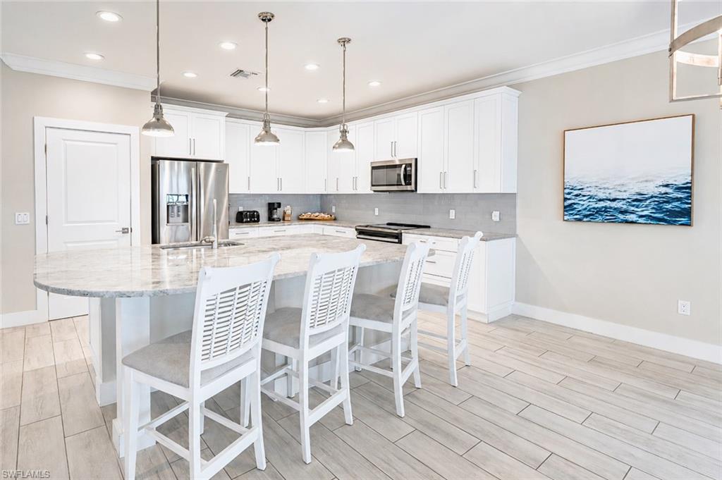 Kitchen featuring a large island with a light-toned countertop, white cabinetry, stainless steel appliances, pendant lighting, and wood-finish flooring - Virtually Edited Image