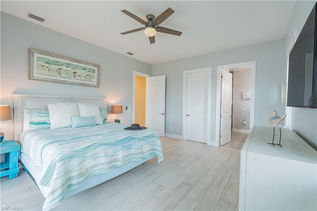 Spacious bedroom featuring light wood-finish flooring, a white paneled headboard, a ceiling fan with integrated lighting, and multiple white interior doors - Virtually Edited Image