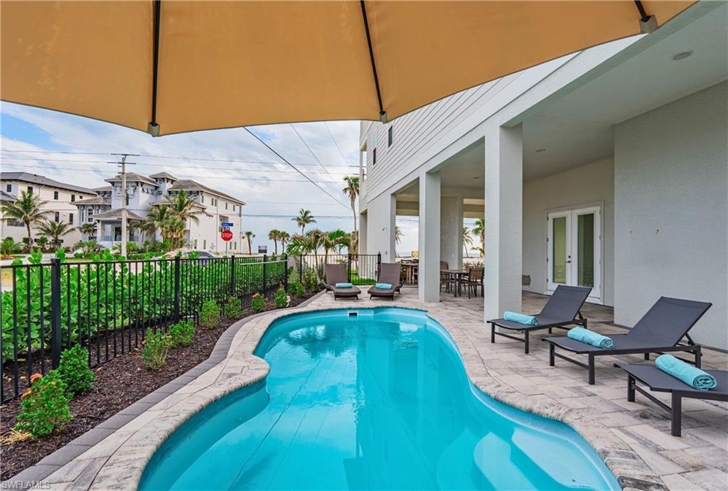 Resort-style pool area featuring a freeform swimming pool, paver patio, covered outdoor living space, and black metal fencing - Virtually Edited Image