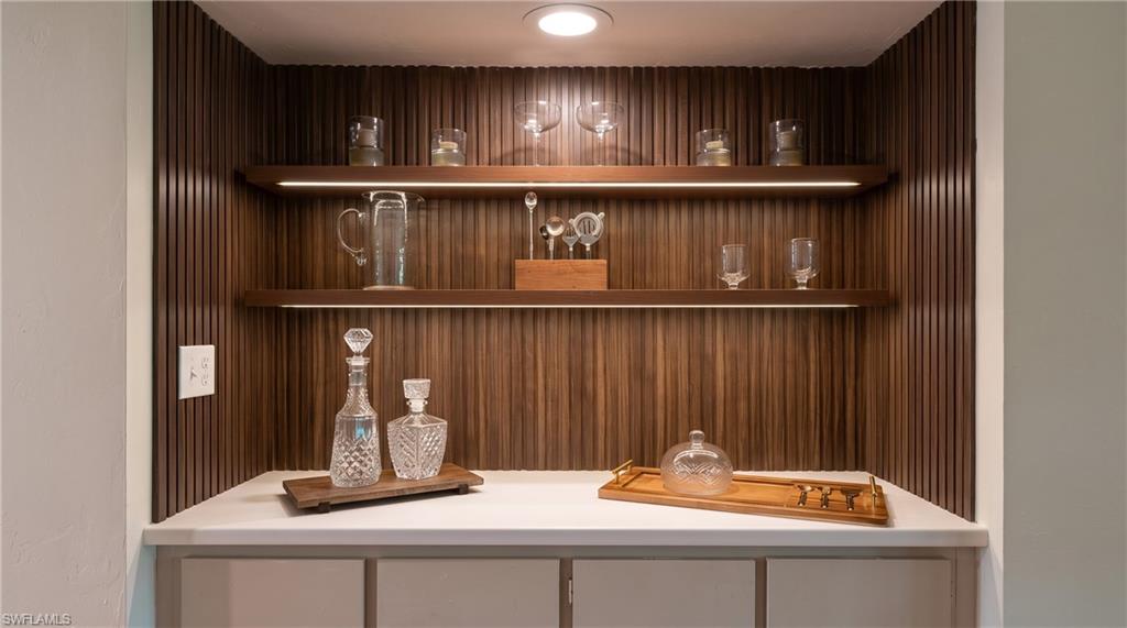Built-in bar niche featuring a slatted wood-finish back panel, two floating shelves with integrated lighting, recessed overhead lighting, and a light-toned countertop above base cabinetry