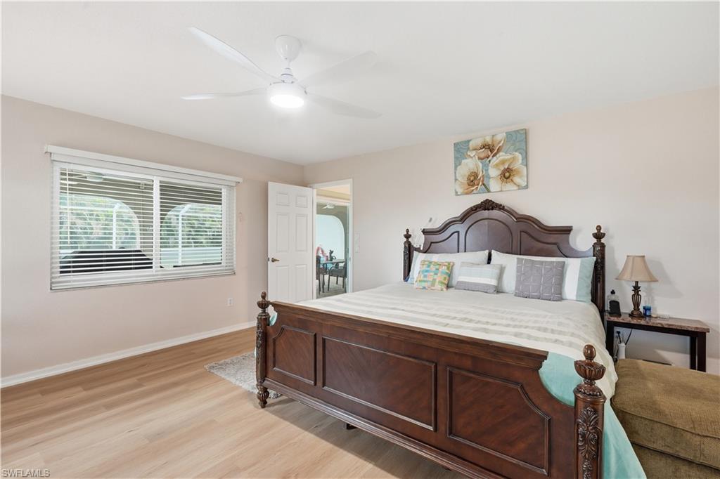 Spacious bedroom featuring light wood-finish flooring, a ceiling fan with integrated lighting, a large window with horizontal blinds, and a white interior door
