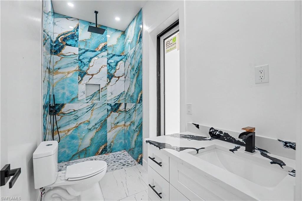 Bathroom featuring a wall-mounted rain shower head, large-format marbled shower tile, recessed shower niche, pebble-style shower floor, and a white vanity with an integrated sink and unique countertop