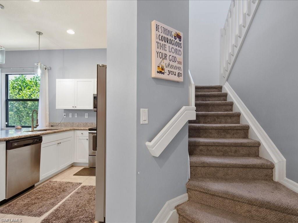 Carpeted staircase with white balustrade and railings, alongside a kitchen featuring white cabinetry, stainless steel appliances, a window, and recessed lighting