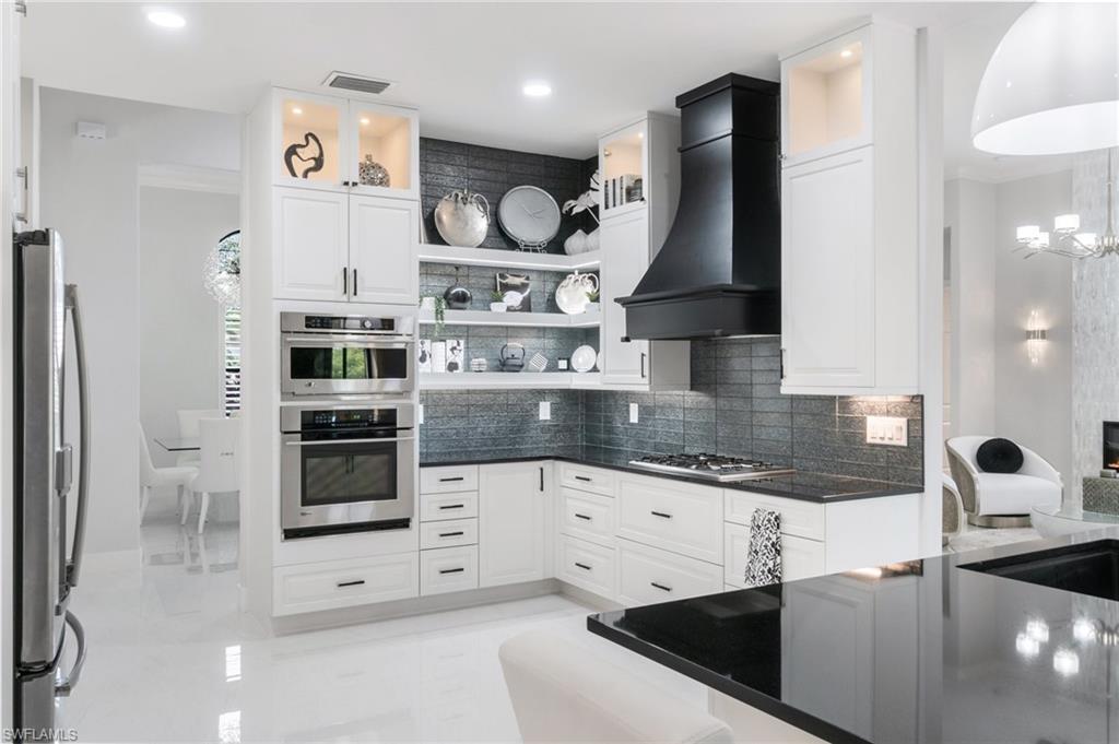 Modern kitchen featuring white cabinetry with black hardware, stainless steel built-in appliances, a black range hood, dark countertops, and a dark tile backsplash