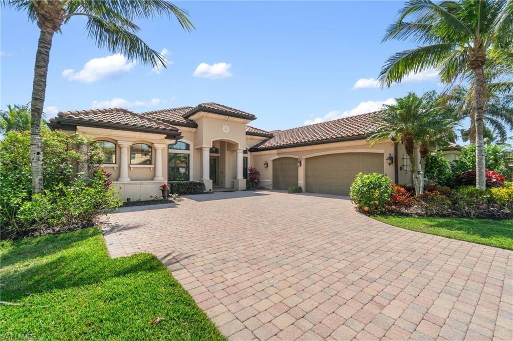 Mediterranean-inspired exterior featuring a paver driveway, tile roof, two-car garage, arched entry, and mature tropical landscaping