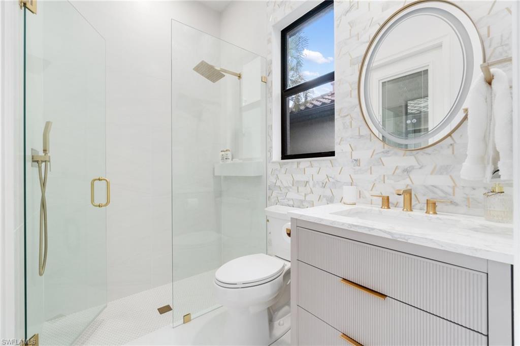 Modern bathroom featuring a frameless glass shower enclosure, a vanity with a white countertop and gold-toned hardware, a circular mirror with integrated lighting, a rain showerhead, and a mosaic tile accent wall