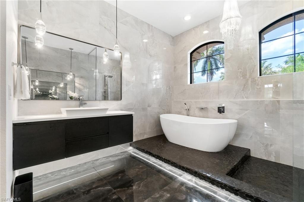 Contemporary bathroom featuring a freestanding soaking tub on a tiered stone platform, dual arched windows, and a floating vanity with vessel sink