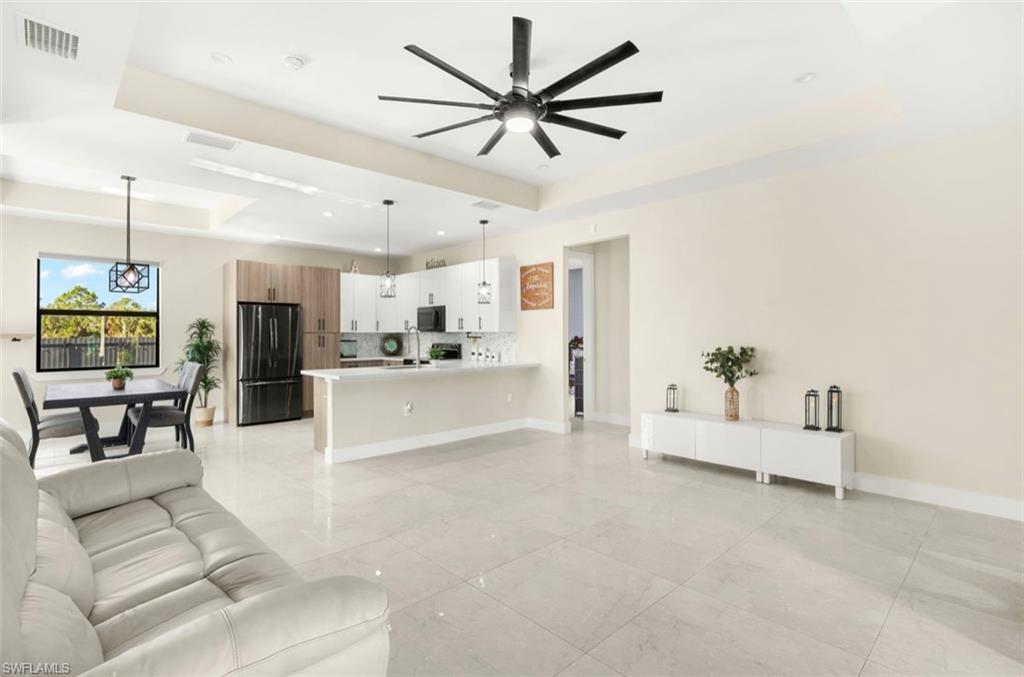 Open-concept living space with large format tile flooring, a contemporary kitchen featuring white cabinetry and black appliances, a dining area with pendant lighting, and a tray ceiling with a modern ceiling fan