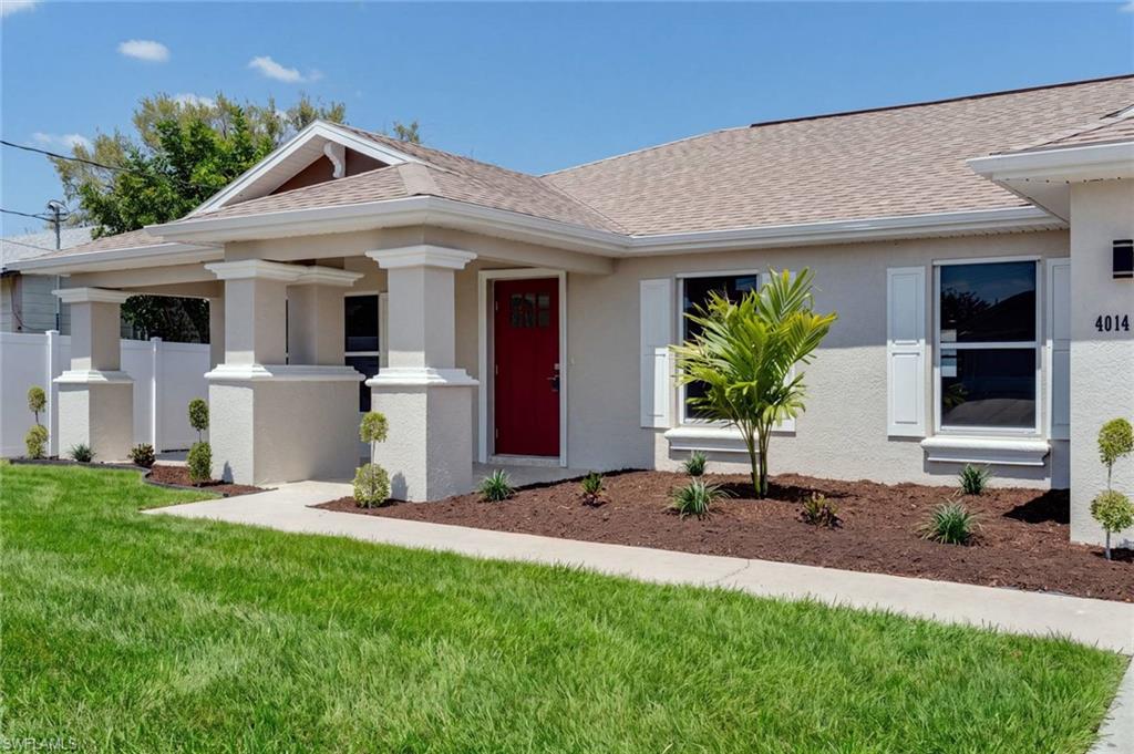 Welcoming front elevation featuring a pitched roofline with architectural columns framing the covered entry, a red lacquered front door, large picture windows with trim detailing, and a manicured lawn with foundational landscaping including a young palm specimen and mulched planting beds