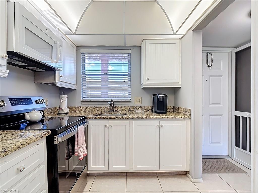 Kitchen with electric stove, white microwave, white cabinets, light stone countertops, and light tile patterned floors