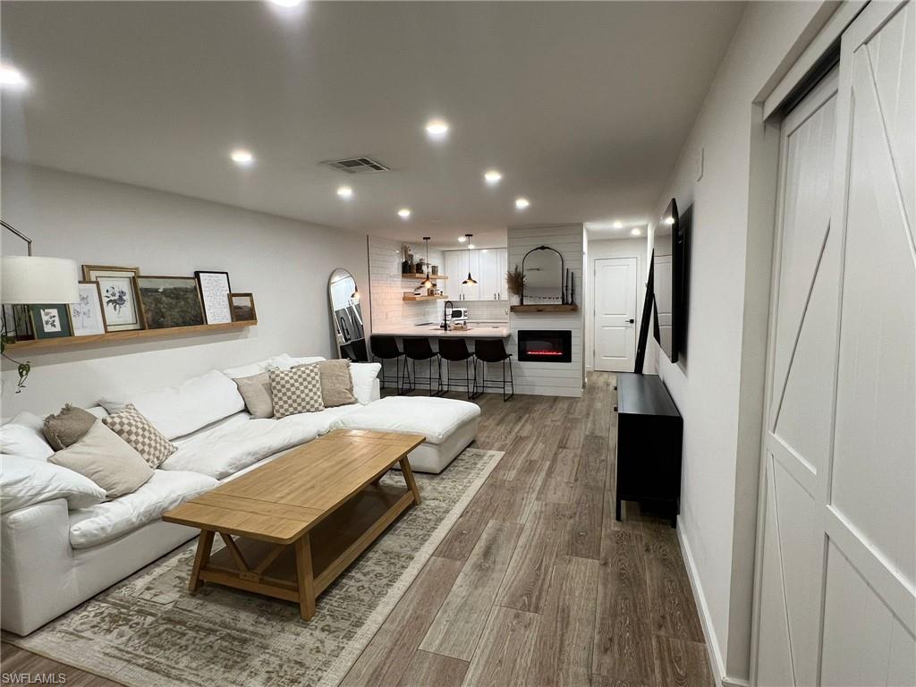 Open-concept living space featuring wood-finish flooring, recessed lighting, a modern electric fireplace with a shiplap surround, and a kitchen island with seating