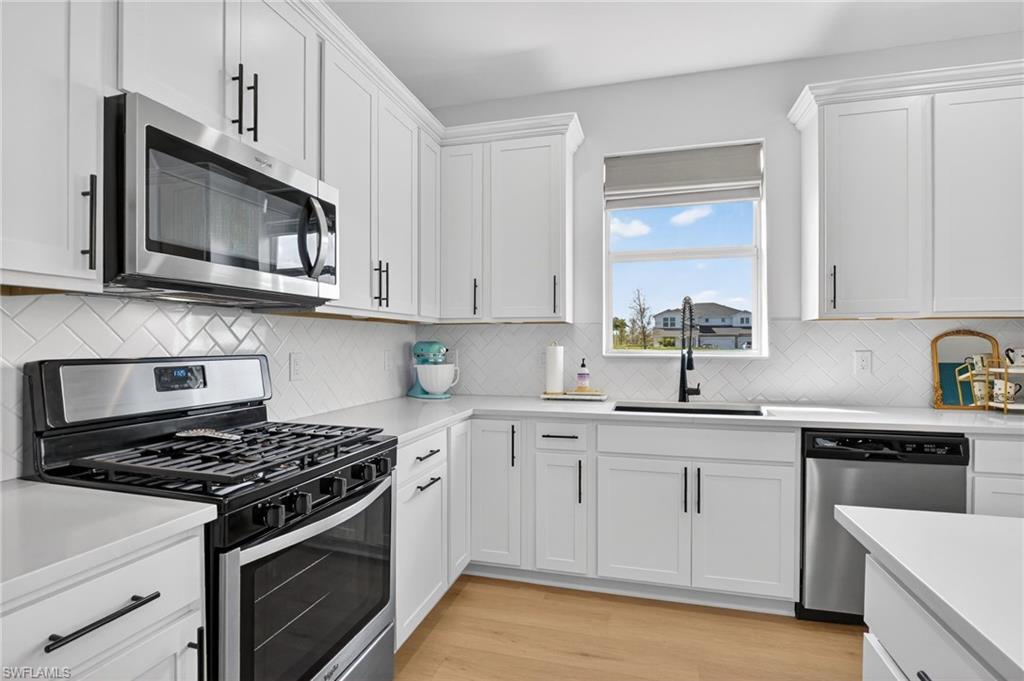 Modern kitchen featuring white cabinetry with black hardware, stainless steel appliances, a white herringbone tile backsplash, light wood-finish flooring, and a window above the sink
