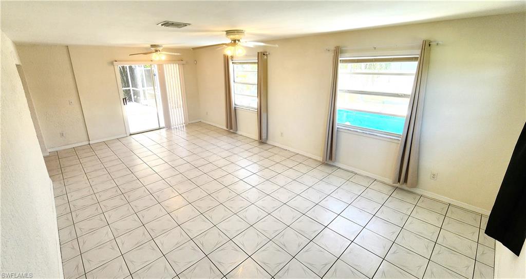 Spacious living area featuring light-toned tile flooring, two ceiling fans, and multiple windows with vertical blinds and drapes