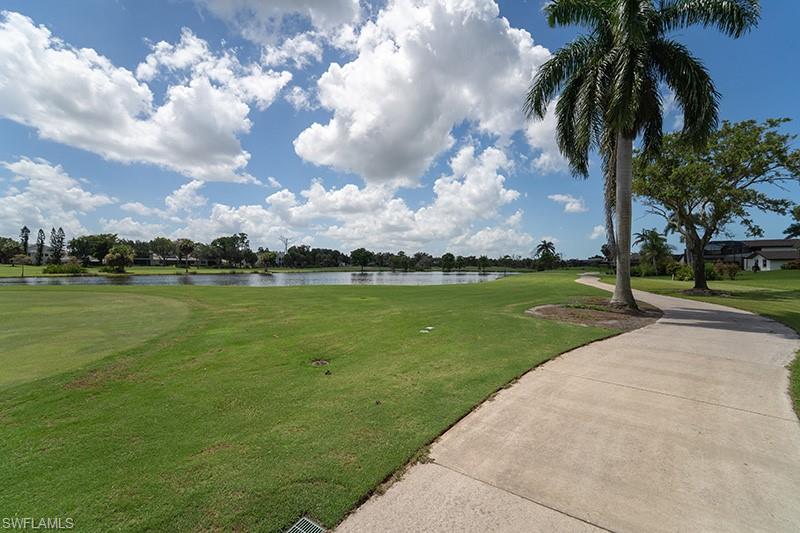 Expansive golf course green with a curving paved pathway, mature palm trees, and a tranquil water feature